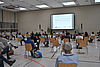 Viele Menschen, die auf Stühlen in einer großen Turnhalle sitzen und in Richtung Vortragenden und Leinwand schauen.