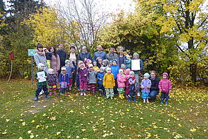 Eine Gruppe von Kindern und Erwachsenen haben sich für ein Gruppenfoto auf einer Wiese mit Herbstlaub und Bäumen im Hintergrund zusammengestellt.