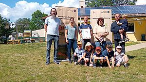 Eine Gruppe von Kindern und Erwachsenen haben sich für ein Gruppenfoto im Grünen auf dem Kitagelände zusammengestellt.