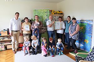 Eine Gruppe von Kindern und Erwachsenen haben sich für ein Gruppenfoto in einem Kitaraum mit Spielzeug im Hintergrund zusammengestellt.