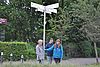 Drei Frauen stehen an einem Hinweisschild für Radfahrwege, im Hintergrund eine grüne Hecke und Laubbäume.