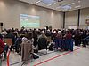 Viele Menschen sitzen in einer großen Halle und schauen auf eine Leinwand.