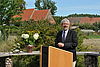Ein Mann im Anzug steht im Freien an einem Rednerpult, neben ihm steht eine Vase mit Blumen, hinter ihm eine Dorfkulisse.