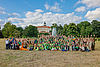 Viele Kinder und einige Erwachsene stehen für ein Gruppenfoto auf einer Wiese zusammen und strecken die Hände nach oben. Im Hintergrund ein Park und ein Schloss.