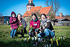 Drei Frauen und ein Kind mit Pflanzen in der Hand auf einer grünen Wiese und vor einer alten Kirche aus Feldsteinen.
