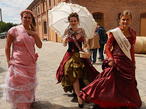Drei Frauen in königlicher Robe, eine mit Sonnenschirm, laufen dem Betrachter entgegen.